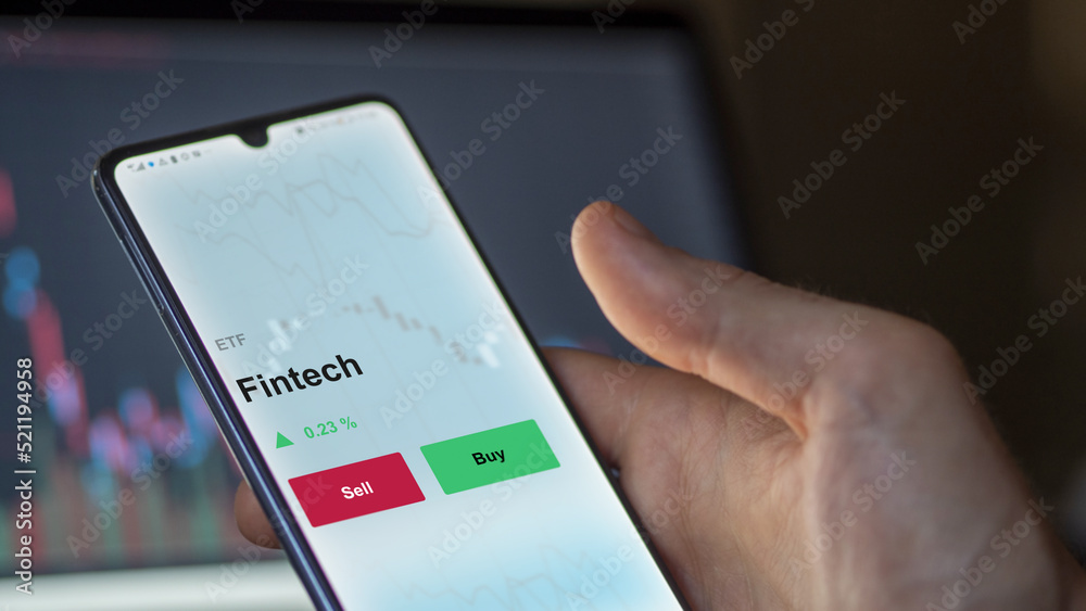 An investor's analyzing the fintech etf fund on a screen. A phone shows the prices of Fintech An investor's analyzing the fintech etf fund on a screen. A phone shows the prices of Fintech