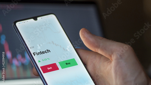 An investor's analyzing the fintech etf fund on a screen. A phone shows the prices of Fintech