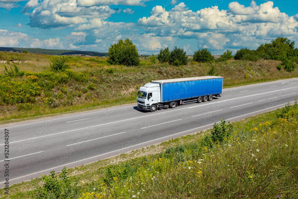 blue cargo truck on the highway. asphalt road among green fields and beautiful clouds. cargo delivery and transportation concept