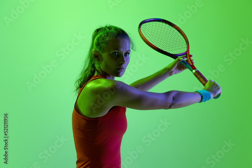 Portrait of young sportive woman, professional tennis player posing isolated over green studio background in neon light. Concentration © Lustre