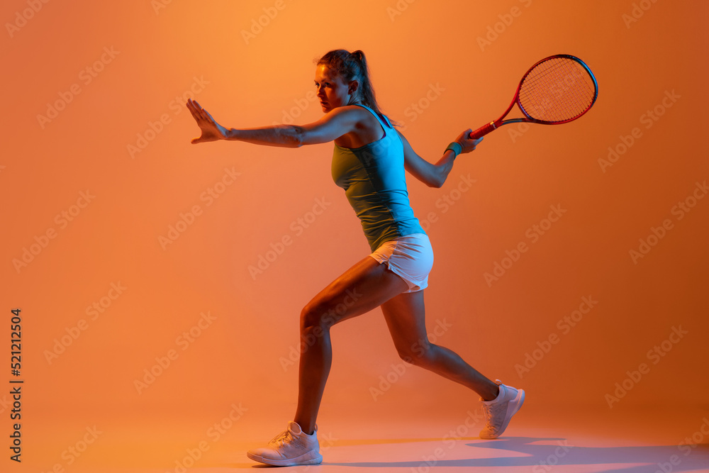 Portrait of concentrated woman, professional tennis player returning ball isolated over orange studio background in neon light