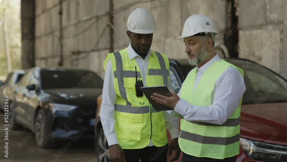Two construction workers in hardhats looking at tablet screen while standing outdoor with automobiles on background. Multiethnic men talking about new architect ideas on working project. Two construction workers in hardhats looking at tablet screen while standing outdoor with automobiles on background. Multiethnic men talking about new architect ideas on working project.