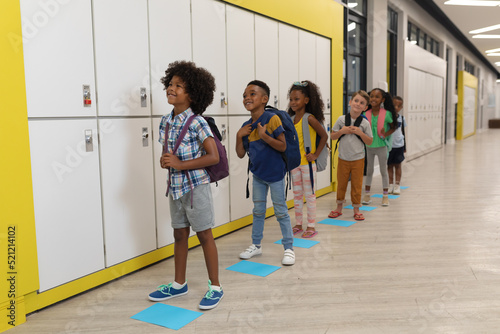Full length of multiracial elementary school children with backpack standing by lockers in corridor
