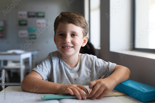 Portrait of smiling caucasian elementary schoolboy with book and pencil sitting at desk in classroom