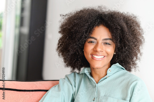 Portrait of beautiful multiracial woman with natural curly afro hairstyle looking at the camera in cozy home atmosphere, charming serene and carefree girl in casual wear smiling toothy