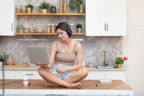 Young beautiful woman in headphones studying online at home. She sits on the table in the kitchen