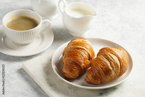 Two french soft croissants on a white plate on a marble tray  a cup of black coffee in white sophisticated cup  milk  sugar  jam