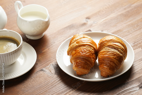 Two french soft croissants on a white plate on a wooden table, a cup of black coffee in white sophisticated cup, milk, sugar, jam