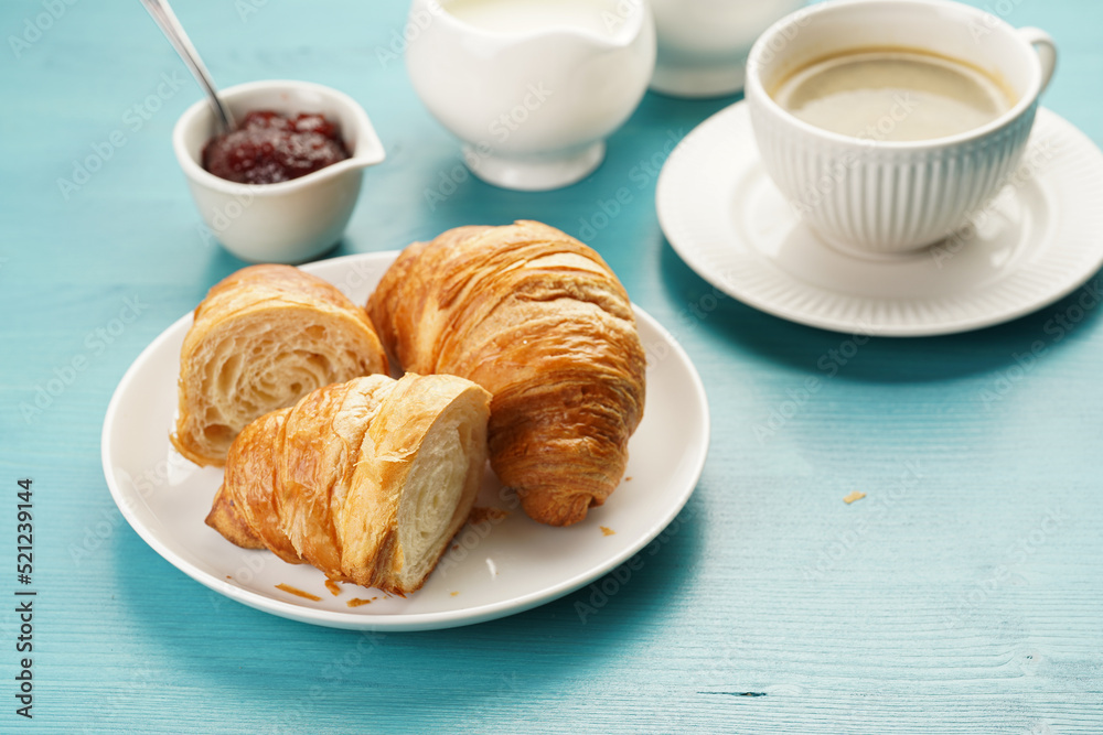 Two french soft croissants on a white plate on a blue wooden table, a cup of black coffee in white sophisticated cup, milk, sugar, jam Two french soft croissants on a white plate on a blue wooden table, a cup of black coffee in white sophisticated cup, milk, sugar, jam