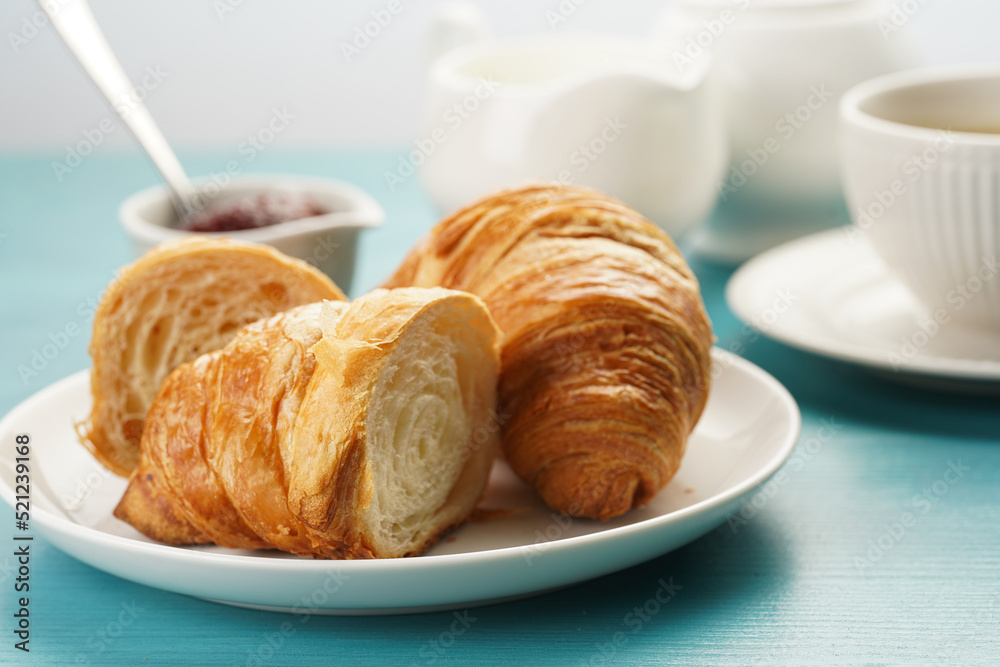 Two french soft croissants on a white plate on a blue wooden table, a cup of black coffee in white sophisticated cup, milk, sugar, jam