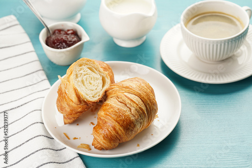 Two french soft croissants on a white plate on a blue wooden table, a cup of black coffee in white sophisticated cup, milk, sugar, jam
