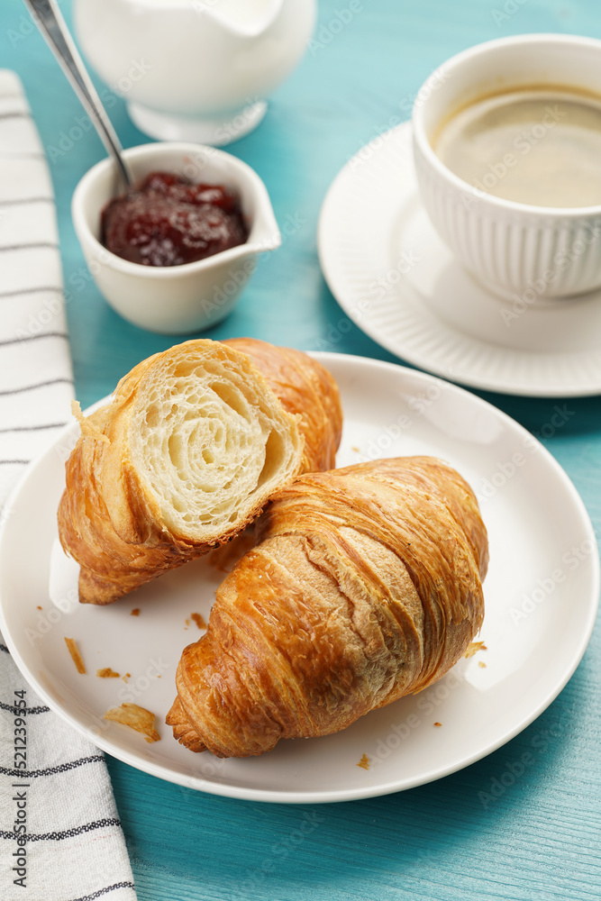 Two french soft croissants on a white plate on a blue wooden table, a cup of black coffee in white sophisticated cup, milk, sugar, jam Two french soft croissants on a white plate on a blue wooden table, a cup of black coffee in white sophisticated cup, milk, sugar, jam