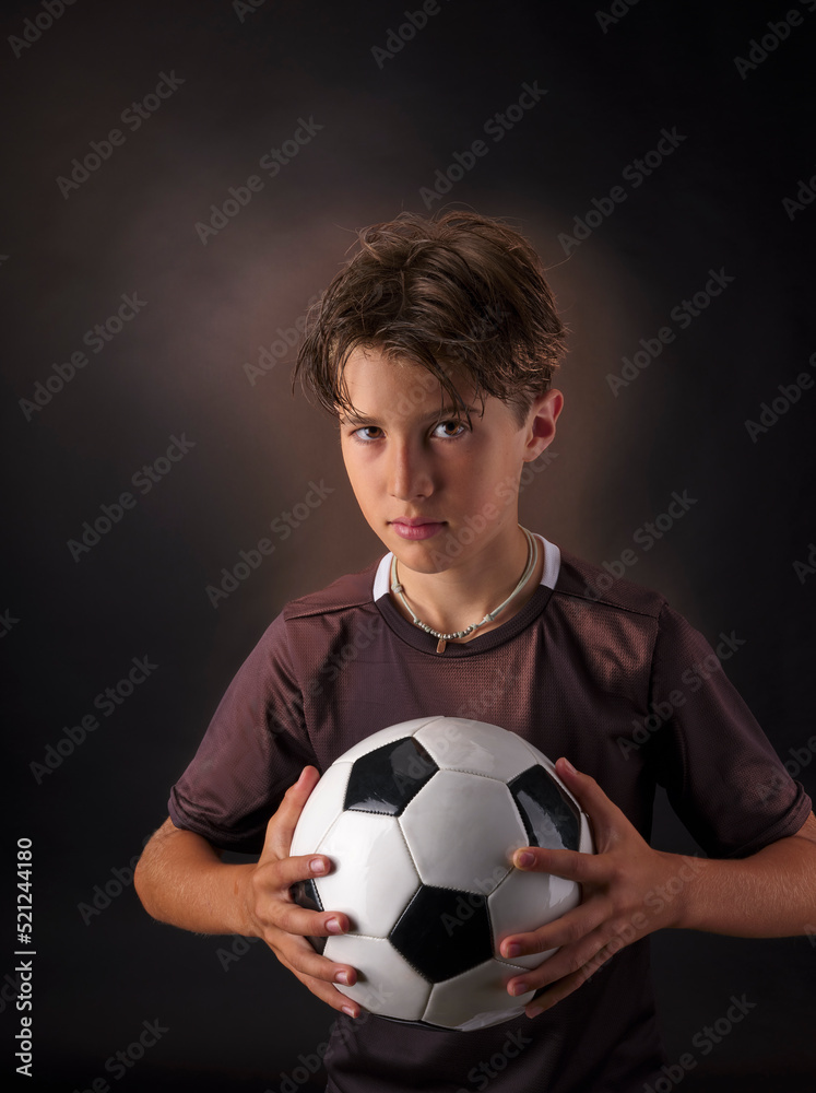 Child posing in the foreground on a black background, with a classic white soccer ball with black pentagon shaped inserts. Child posing in the foreground on a black background, with a classic white soccer ball with black pentagon shaped inserts.