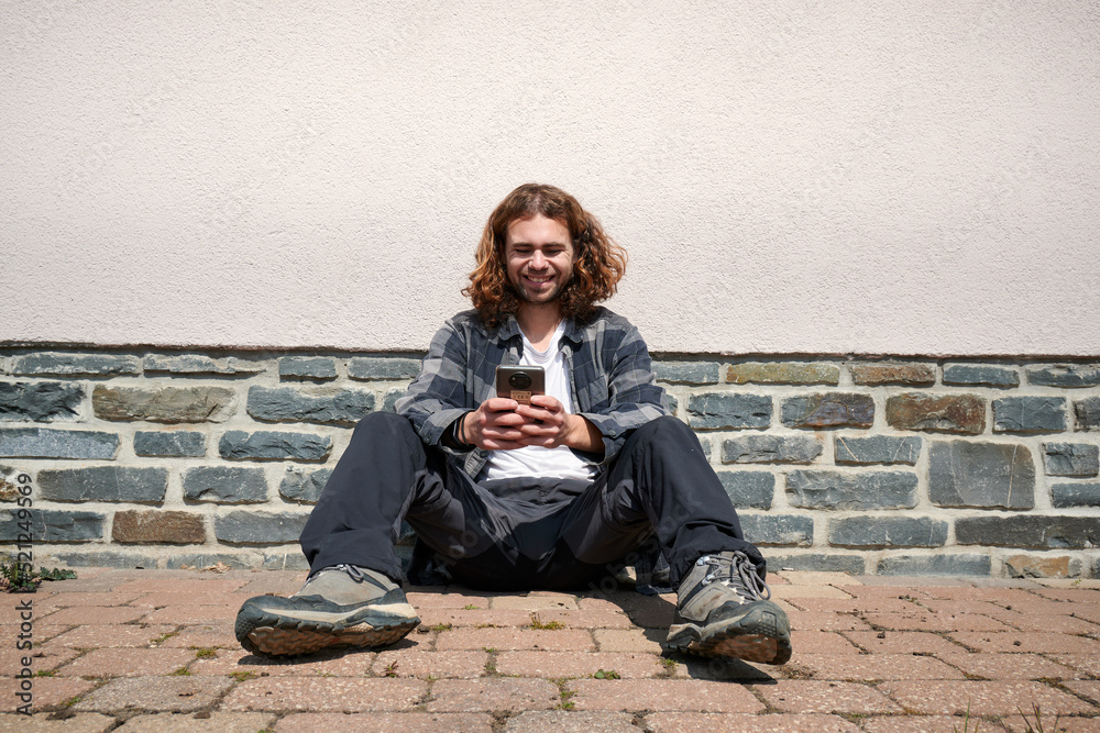 Fototapeta premium A happy young Caucasian guy sitting in his phone against the wall
