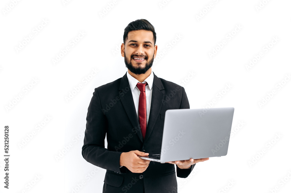 Positive elegant attractive indian or arabian man in business suit, entrepreneur, holding open laptop in hands, looks at camera, standing over isolated white background, smiles friendly. Copy-space Positive elegant attractive indian or arabian man in business suit, entrepreneur, holding open laptop in hands, looks at camera, standing over isolated white background, smiles friendly. Copy-space