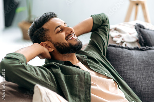 Relaxed satisfied calm mixed race guy in casual shirt, resting on the sofa in cozy living room with his eyes closed, throwing his hands behind his head, dreaming of vacation, resting from work, smiles