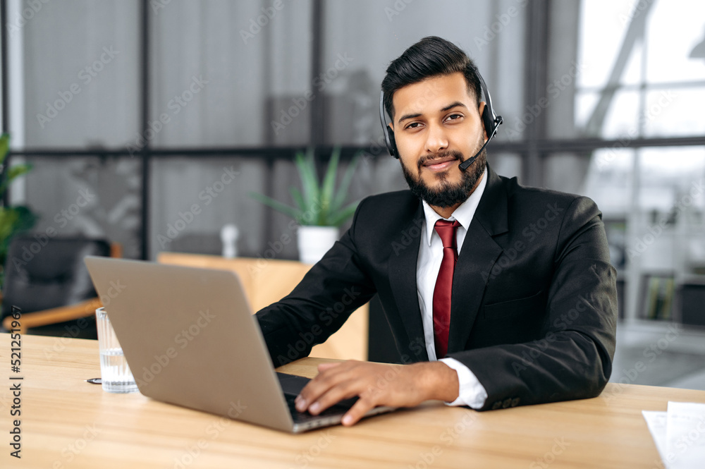 Confident clever indian or arabian young man in suit and with headset, business advisor, call center worker, company representative, support service operator, holds online consultation,looks at camera