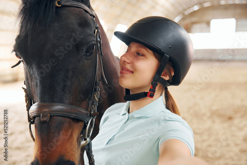 Making lovely selfie. A young woman in jockey clothes is preparing for a ride with a horse on a stable © standret