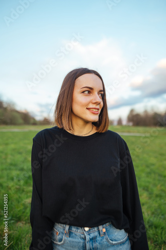 Vertical portrait of a smiling attractive woman in casual clothes standing in a park at sunset and looking away with a smile on her face.