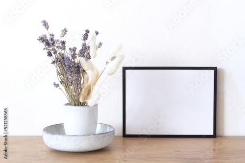 Horizontal black frame mockup on a vintage wooden bench, table. Modern white ceramic vase with dry Lagurus ovatus grass and lavender. White wall background. Scandinavian interior. Selective focus.