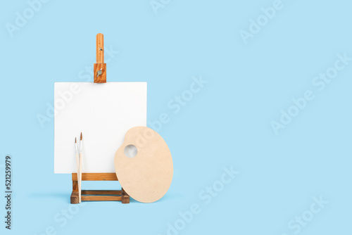 Leinwand Poster Lienzo en blanco con caballete de madera junto a una paleta y pinceles sobre un fondo celeste claro liso y aislado