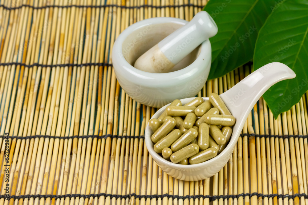 Mitragyna Speciosa Korth or kratom capsules on white bowl with green leaf on wooden mat. 