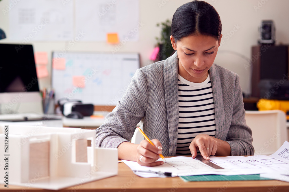 Female Architect Working In Office With Model On Desk Studying Plans For New Building