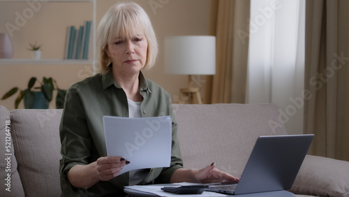 Caucasian old 50s homeowner housewife makes woman budget checks monthly income by checking paper notes using app laptop. Middle-aged businesswoman sitting on couch with document working remote at home
