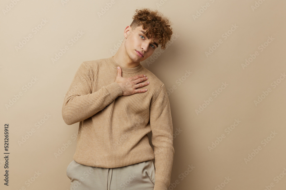 a charming man in a beige sweater stands on a beige background and it's nice to look into the camera with his hand on his body a charming man in a beige sweater stands on a beige background and it's nice to look into the camera with his hand on his body