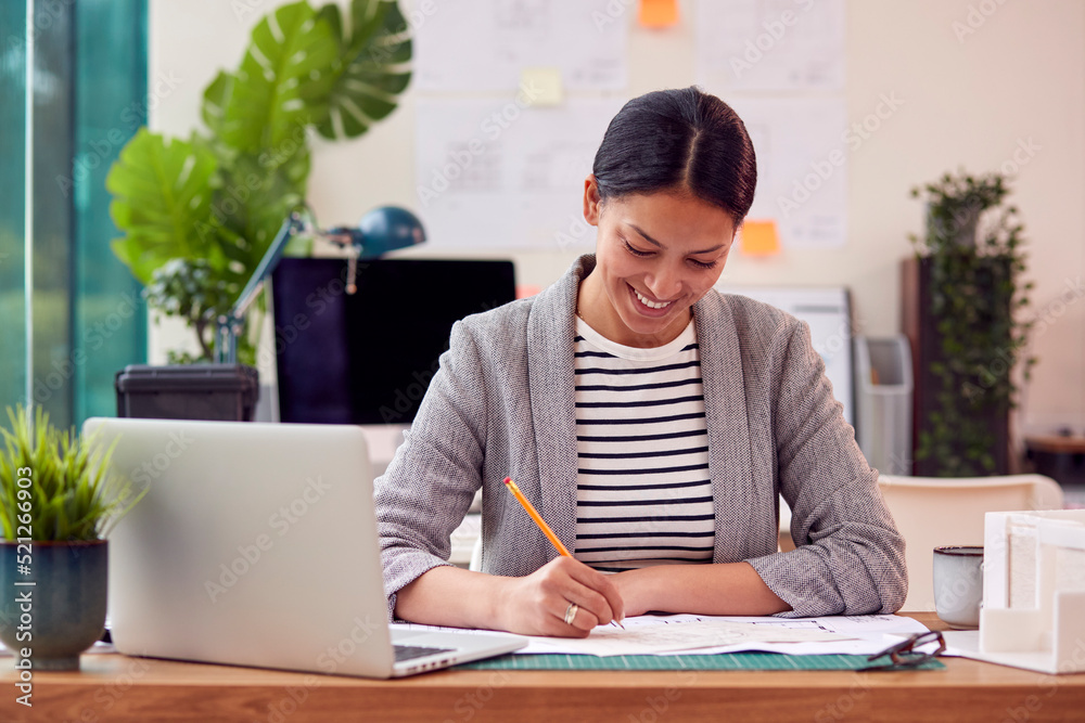 Female Architect Working In Office Sitting At Desk Studying Plans For New Building Female Architect Working In Office Sitting At Desk Studying Plans For New Building