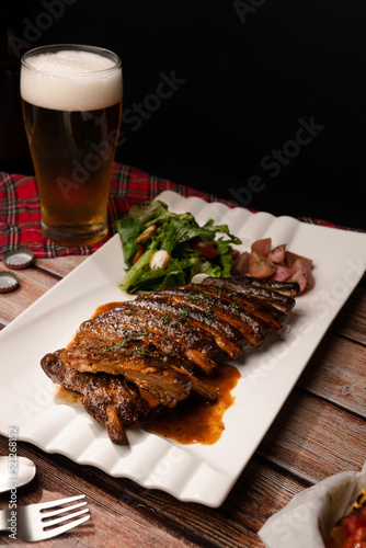 Close up grilled bbq ribs serve in white square plate with glass of beer on wood table