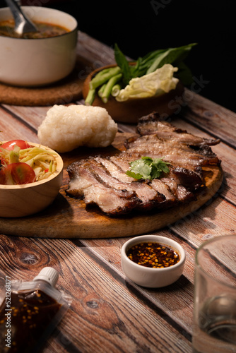 Close up Thai food Grilled Pork jowl serve on round wood plate with spicy papaya salad sticky rice spicy sauce on wood table with drink snack and appetizer meal