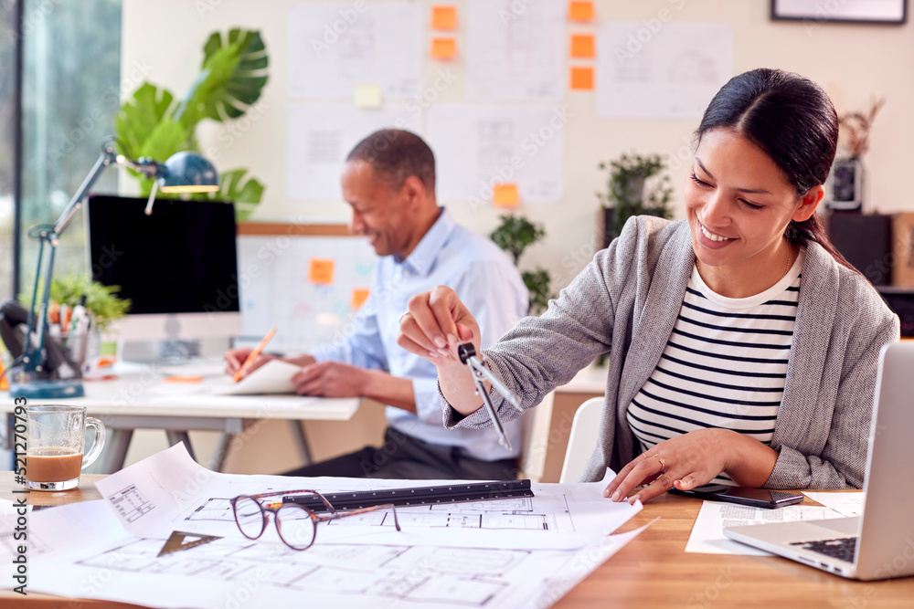 Male And Female Architects Working In Office At Desks On Plans For New Building Male And Female Architects Working In Office At Desks On Plans For New Building