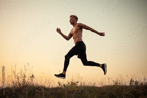 Athletic young guy running on grassy meadow under sundown sky