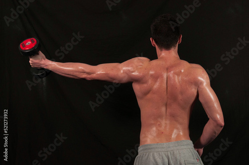 Back view of a shirtless young bodybuilding athlete doing dumbbell lateral raises in a gym. photo