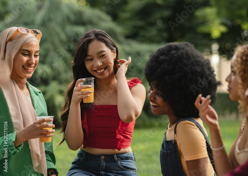 multiracial female friends having drink in park - focus on asian woman -
