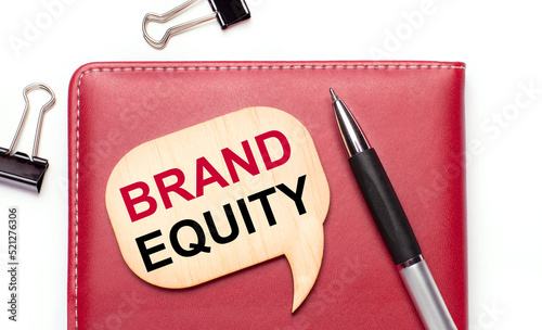 On a light background there are black paper clips, a pen, a burgundy notepad a wooden board with the text BRAND EQUITY
