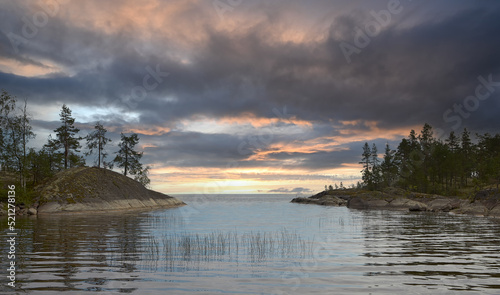 Evening landscape. Summer. Lake Ladoga. In the foreground is a strait surrounded by rocky shores. Dark gray sky with reflections of the setting sun.