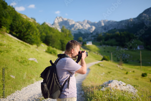 Mature man tourist with backpack and camera is hiking on mountain valley. Photographer taking a picture during travel.
