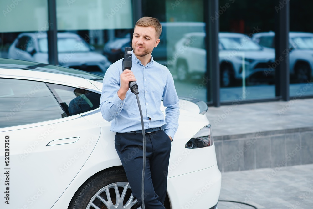 Full length portrait of young handsome bearded man in casual wear, standing at the charging station and holding a plug of the charger for an electric car. Eco electric car concept.