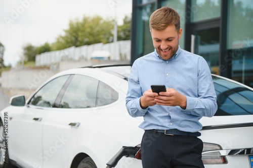 Handsome man in business suit surfing internet on modern smartphone while waiting electric car to charge.