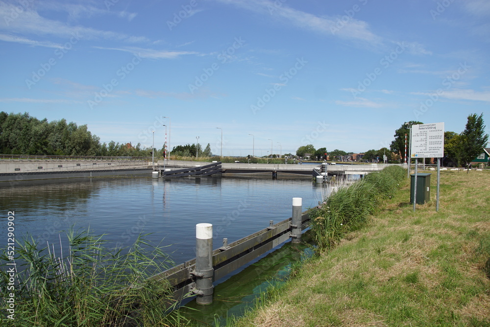 New float bridge Rekervlotbrug with cycle path in a canal in the Dutch village of Koedijk. Retractable pontoon bridge which opens for water traffic by retracting. Board with operating times. New float bridge Rekervlotbrug with cycle path in a canal in the Dutch village of Koedijk. Retractable pontoon bridge which opens for water traffic by retracting. Board with operating times.