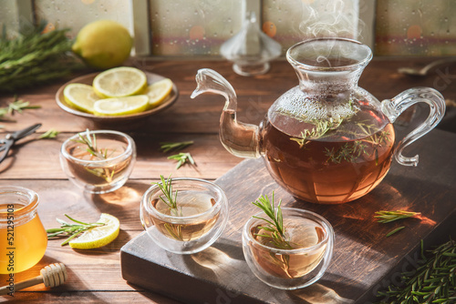 Hot rosemary tea in glass teapot and cups. Served with honey and fresh lemon on rustic wooden background. 