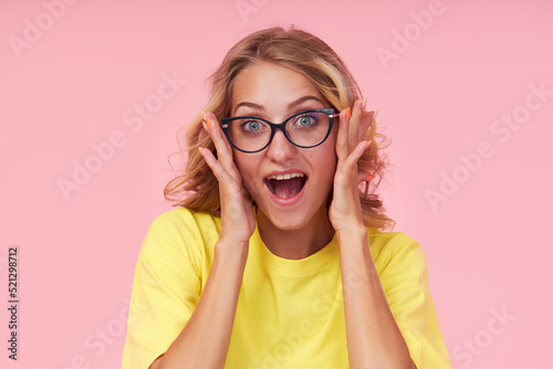 Portrait of an excited young woman in eyeglasses, yellow t-shirt on a pink background. Surprised girl looks to the right with her eyes wide open. Wow face