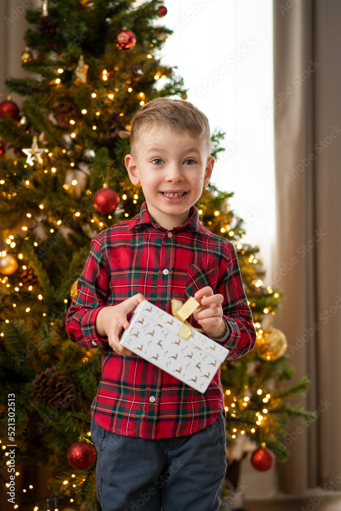 A little smiling boy stands next to the Christmas tree and opens a box with a gift
