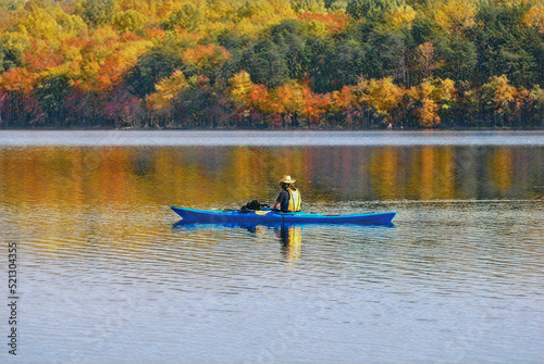 Kayaking Burke Lake in Virginia, with fall foliage.  A person, a man, with a yellow life jacket and cowboy hat is in the blue kayak. 