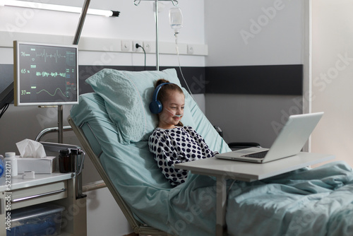 Happy sick little girl under treatment sitting on patient bed with laptop while watching cartoons inside hospital pediatrics ward room. Ill kid having oxygen tube enjoying funny videos on computer. photo