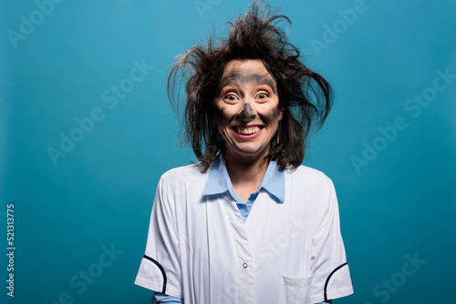 Portrait of crazy chemist with dirty face and messy hair grinning dreadful at camera while on blue background. Mad scientist with dreadful smile and stupid look after chemical laboratory explosion.