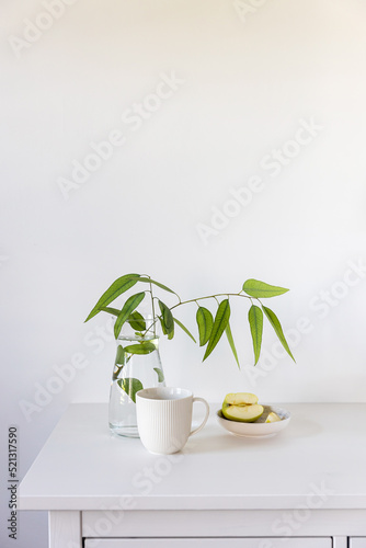 eucalyptus branch in a glass vase on a white table, a sliced green apple in a saucer against the background of the wall. Ready layout. Vertical frame. Space for text
