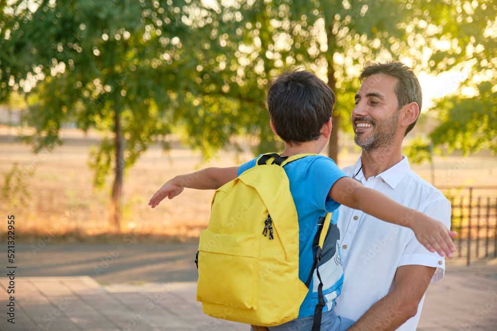 smiling dark-haired father in white shirt holding child in arms in blue t-shirt and yellow backpack smiling dark-haired father in white shirt holding child in arms in blue t-shirt and yellow backpack